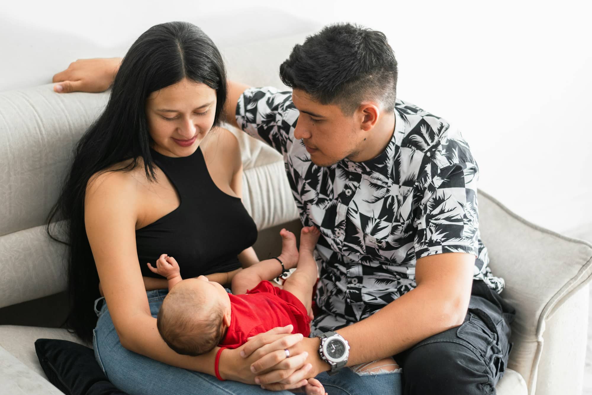 couple of young Latino parents sitting on the couch while proudly looking at their baby girl